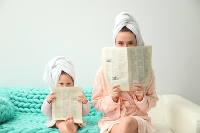 Mother and little daughter in bathrobes with newspapers sitting on sofa