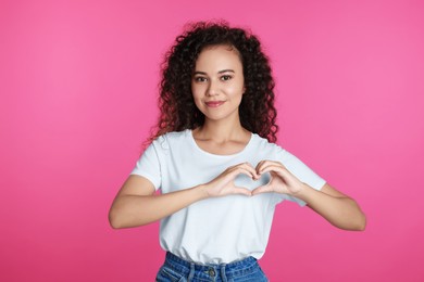 Happy young African-American woman making heart with hands on pink background