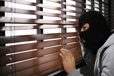 Masked man spying through window blinds indoors. Criminal offence