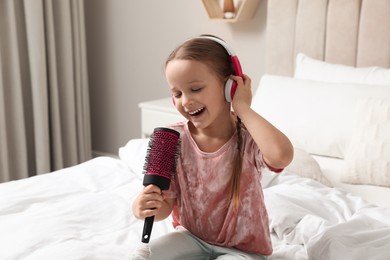 Cute little girl in headphones with hairbrush singing on bed at home