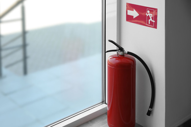 Modern fire extinguisher and emergency exit sign near window indoors. Space for text