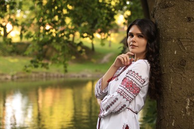 Beautiful woman in embroidered shirt near lake, space for text. Ukrainian national clothes