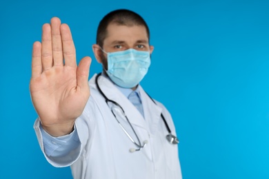 Doctor in protective mask showing stop gesture on light blue background, space for text. Prevent spreading of coronavirus