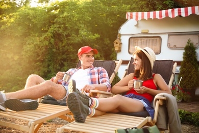Happy couple with cups resting on deck chairs near motorhome. Camping season