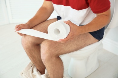 Young man with bath tissue sitting on toilet bowl at home