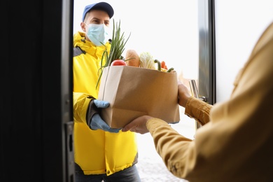 Courier in medical mask giving paper bag with groceries to woman at doorway. Delivery service during Covid-19 quarantine