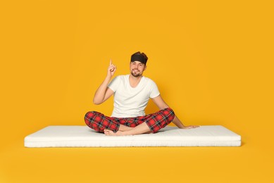 Smiling man in sleeping mask sitting on soft mattress and pointing upwards against orange background