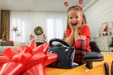 Excited little girl sitting inside toy car in room decorated for Christmas