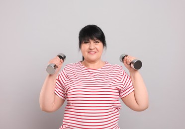 Happy overweight mature woman doing exercise with dumbbells on grey background
