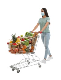 Young woman in medical mask with shopping cart full of groceries on white background