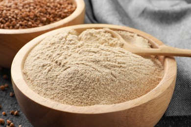 Buckwheat flour in bowl with wooden spoon on black table, closeup