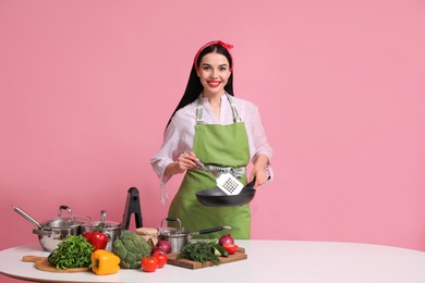 Young housewife with pan, spatula and products on pink background