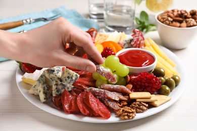 Woman taking sausage from plate with different appetizers on white wooden table, closeup