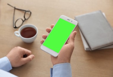 Image of Man holding smartphone with green screen at wooden table indoors, closeup. Gadget display with chroma key. Mockup for design