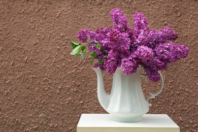 Beautiful lilac flowers in teapot near brown wall. Space for text