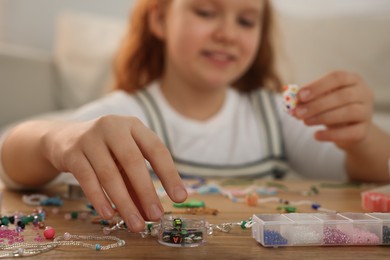 Cute girl making beaded jewelry at table in room, focus on hand