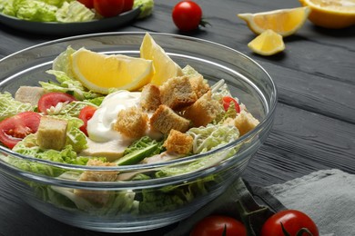 Photo of Bowl of delicious salad with Chinese cabbage, lemon, tomatoes and bread croutons on black wooden table, closeup