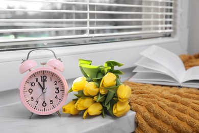 Photo of Pink alarm clock, soft blanket, book and wonderful tulips on window sill indoors. Spring atmosphere