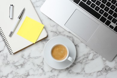 Notebook with blank sticky note, laptop and cup of coffee on white marble table, flat lay