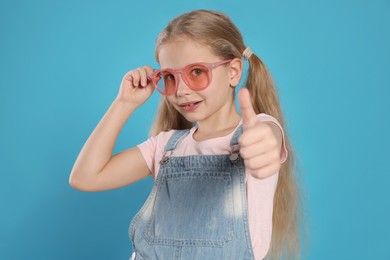Girl in pink sunglasses showing thumb up on light blue background
