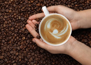 Image of Woman with cup of tasty cappuccino and roasted coffee beans, top view