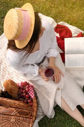 Woman with glass of wine, book and picnic basket on green grass, above view