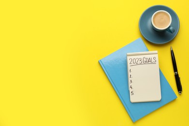 Inscription 2023 Goals written in notebook, pen and coffee on yellow background, flat lay with space for text. New Year aims