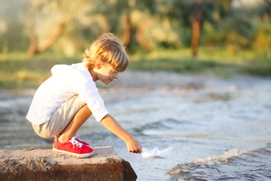 Photo of Cute little boy with paper ship at river, space for text. Child spending time in nature