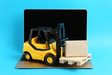 Laptop, toy forklift with wooden pallet and box on light blue background