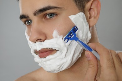 Handsome young man shaving with razor on grey background, closeup