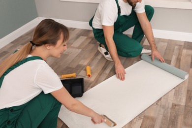 Workers applying glue onto wall paper sheet on floor indoors