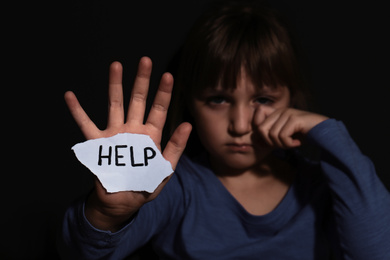 Crying little girl with sign HELP near black wall, focus on hand. Domestic violence concept