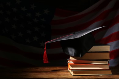 Graduation hat and books on wooden table against American flag in darkness, space for text