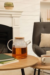 Teapot, cups of hot drink and books on wooden tables near armchair in room. Interior design