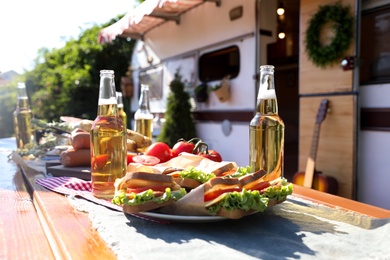 Delicious sandwiches and bottles of beer on wooden table near motorhome on sunny day. Camping season