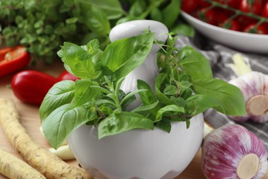 Photo of Mortar with different fresh herbs near garlic and horseradish roots on table, closeup