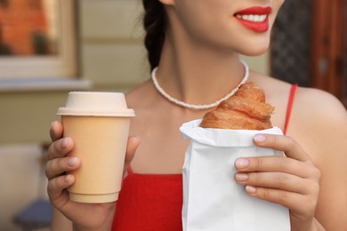 Woman with croissant and paper cup of coffee outdoors, closeup