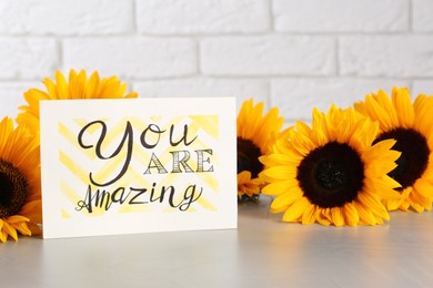 Card with life-affirming phrase You Are Amazing and sunflowers on light table against white brick wall