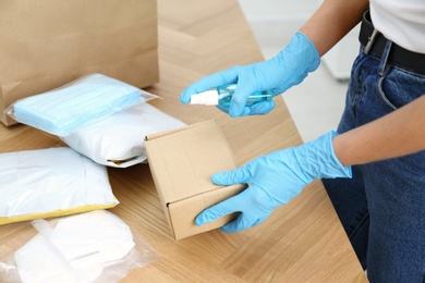 Woman spraying antiseptic onto parcel at wooden table, closeup. Preventive measure during COVID-19 pandemic
