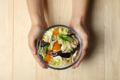 Photo of Woman holding bowl of delicious salad with Chinese cabbage, tomato and basil at wooden table, top view