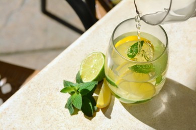 Pouring water into glass with lemon slices and mint from jug at light grey table, closeup. Space for text