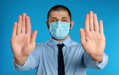 Man in protective mask showing stop gesture on light blue background. Prevent spreading of coronavirus
