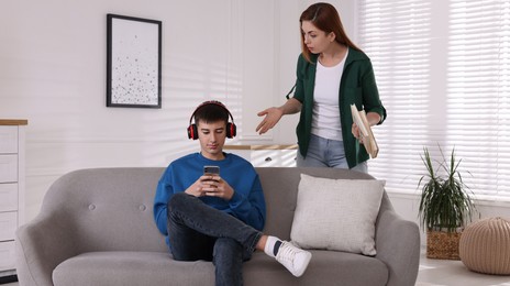 Strict mother with books scolding her son while he using smartphone at home. Teenager problems