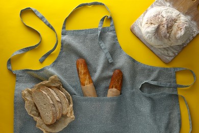 Clean kitchen apron with different types of bread on yellow background, flat lay
