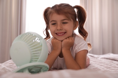 Little girl enjoying air flow from portable fan on bed in room. Summer heat