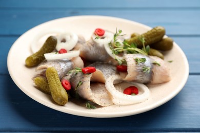 Photo of Plate with salted herring fillets, onion rings, thyme, pickles and chili pepper on blue wooden table, closeup