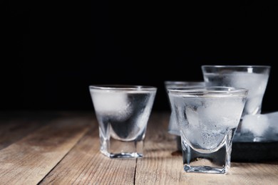 Photo of Shot glasses of vodka with ice cubes on wooden table against black background, closeup. Space for text