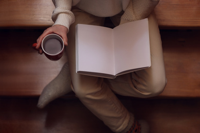 Woman with cup of coffee reading book on stairs at home, top view