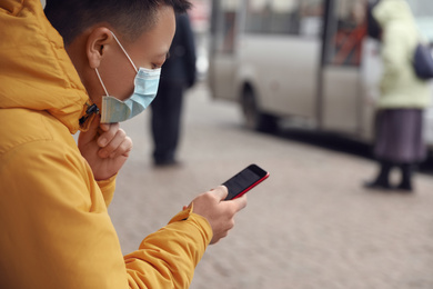 Asian man with smartphone wearing medical mask on city street, space for text. Virus outbreak