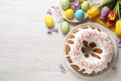 Glazed Easter cake with sprinkles, painted eggs and flowers on white wooden table, flat lay. Space for text